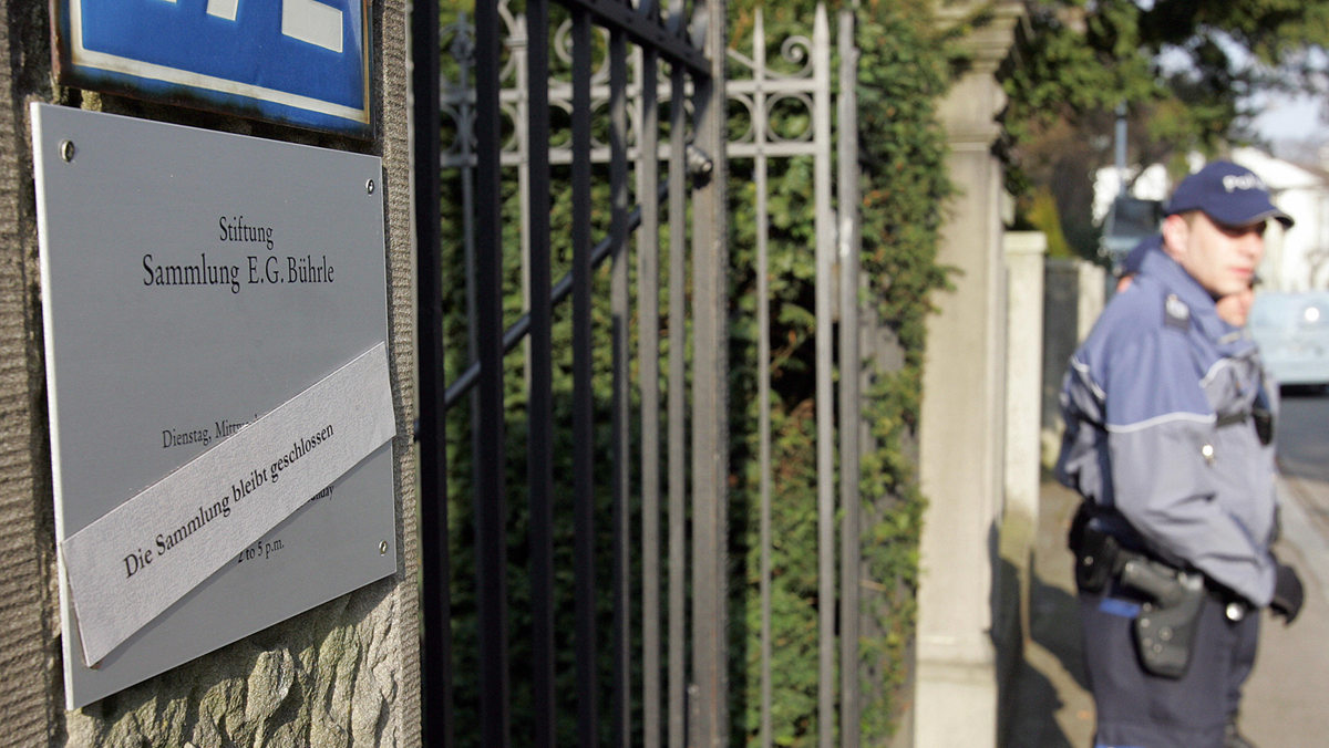 A police officer stands near a metal gate with a sign that reads “Stiftung Sammlung E.G. Bührle.” The sign is partially covered by police tape. Trees and buildings are visible in the background.