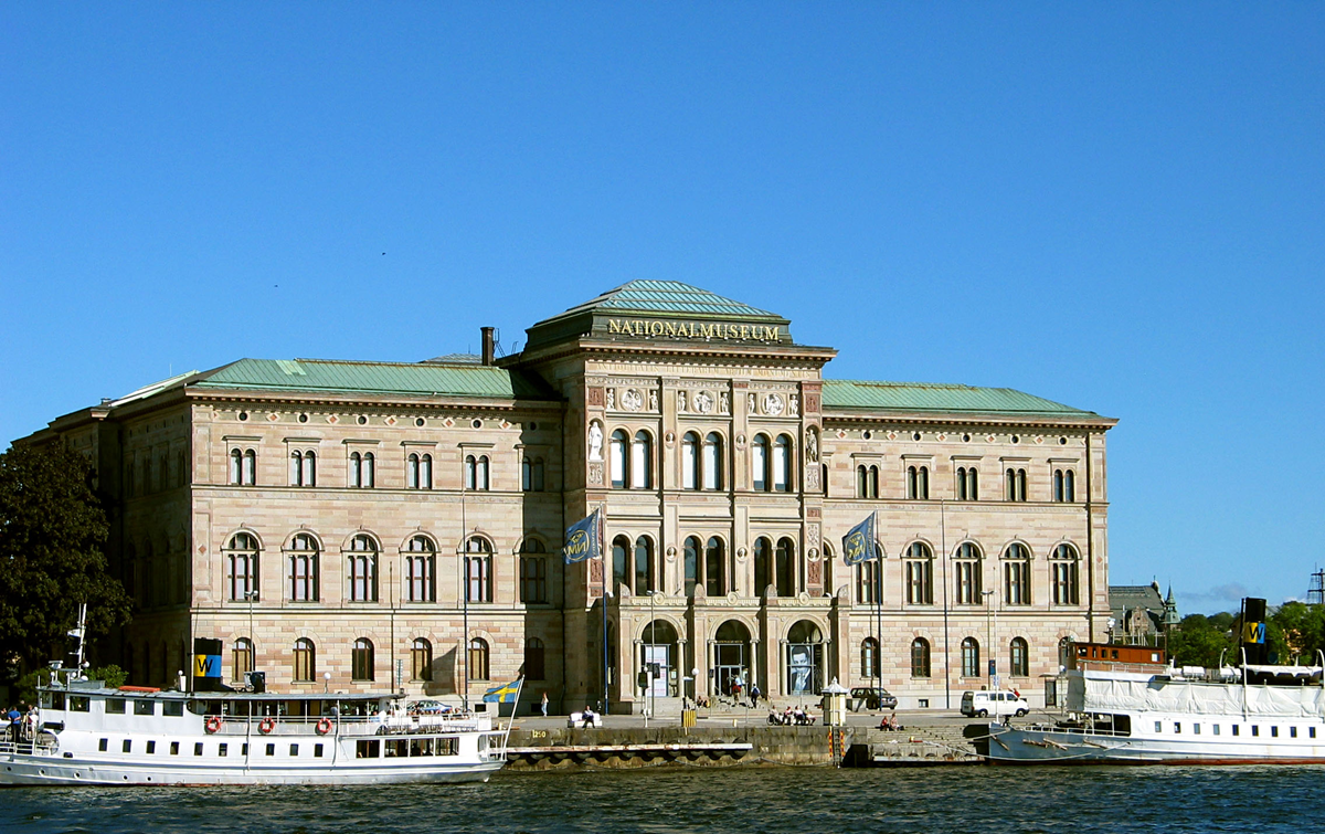 Large, historic stone building with arched windows beside water, boats docked in front, flags flying, clear blue sky above.