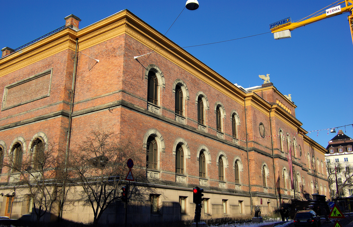 A large brick building with arched windows, stone trim, and a yellow crane nearby, under a clear blue sky. Bare trees and a traffic light are visible in the foreground.