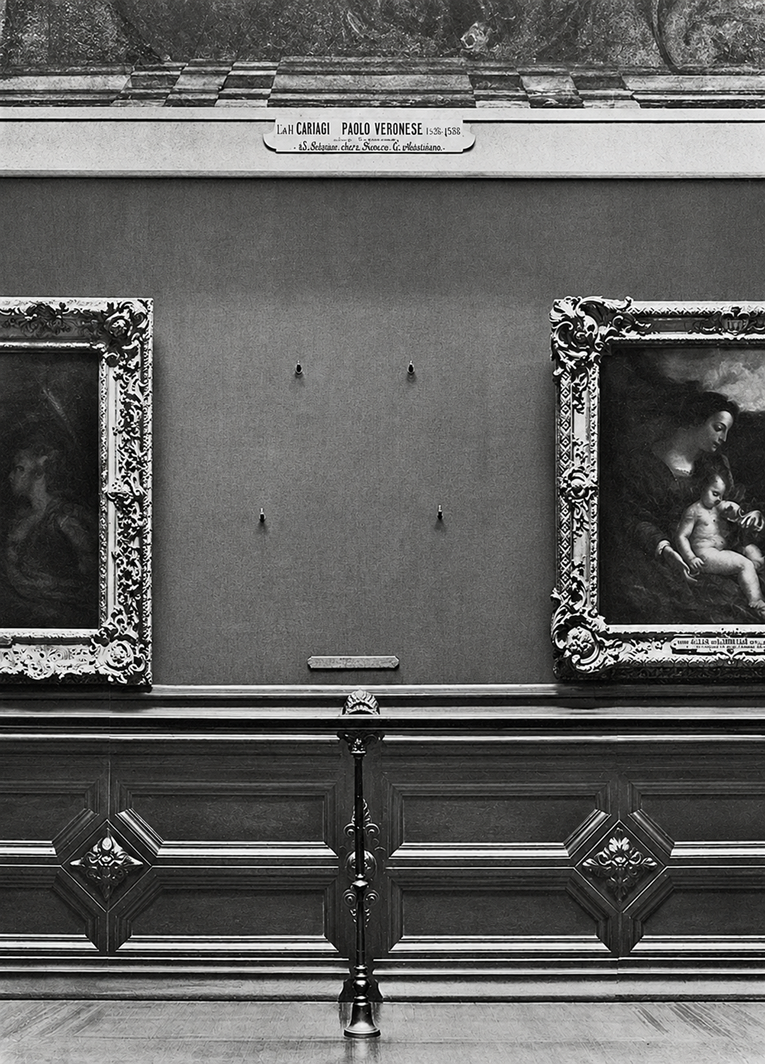 A museum wall displays two framed paintings with an empty space between them, marked by four hooks and a nameplate above reading “Paolo Veronese.” The area is roped off, and ornate wood paneling lines the lower wall.