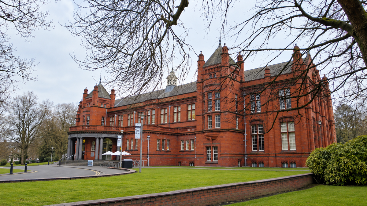 A large historic red-brick building with ornate architecture, arched windows, and a columned entrance, surrounded by green lawns, trees, and a pathway.
