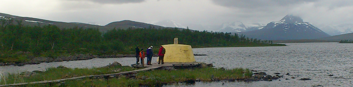 Four people stand by a yellow structure on a lakeshore dock surrounded by rocks and greenery, with cloudy skies and snow-capped mountains in the background.