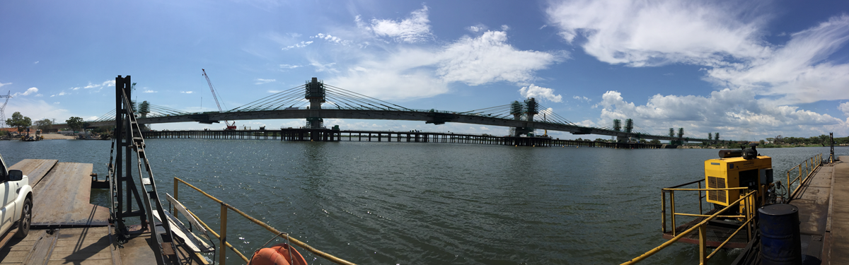 A wide cable-stayed bridge under construction spans a river, with cranes and scaffolding visible. Blue sky with scattered clouds is above, and construction equipment lines the waterfront.