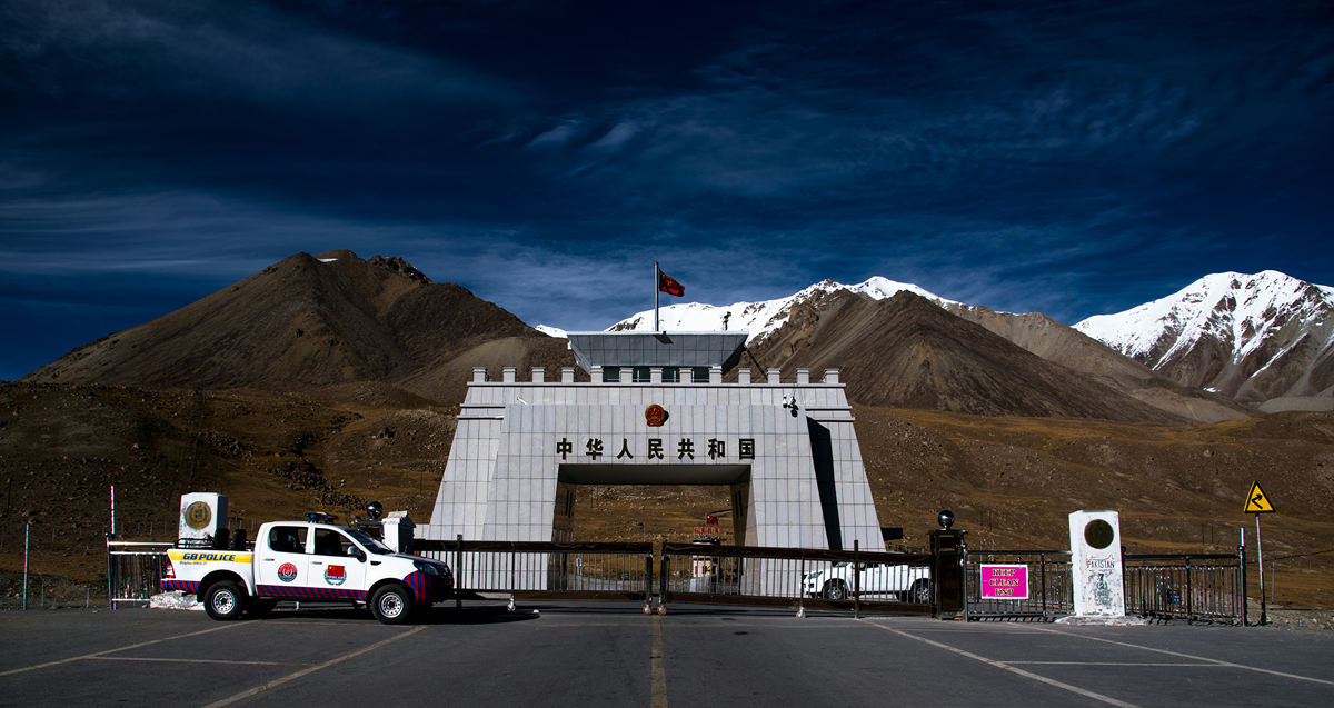 A white gatehouse at a mountain border crossing under a deep blue sky, with a Chinese flag atop, mountains in the background, and a white vehicle parked at the closed gate.