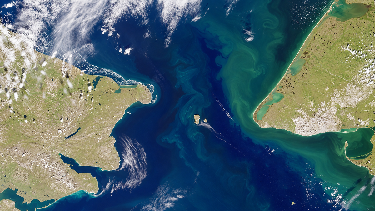 Satellite view of the Bering Strait, showing swirling greenish-blue ocean currents between Russia and Alaska, with clouds scattered above the land and water.