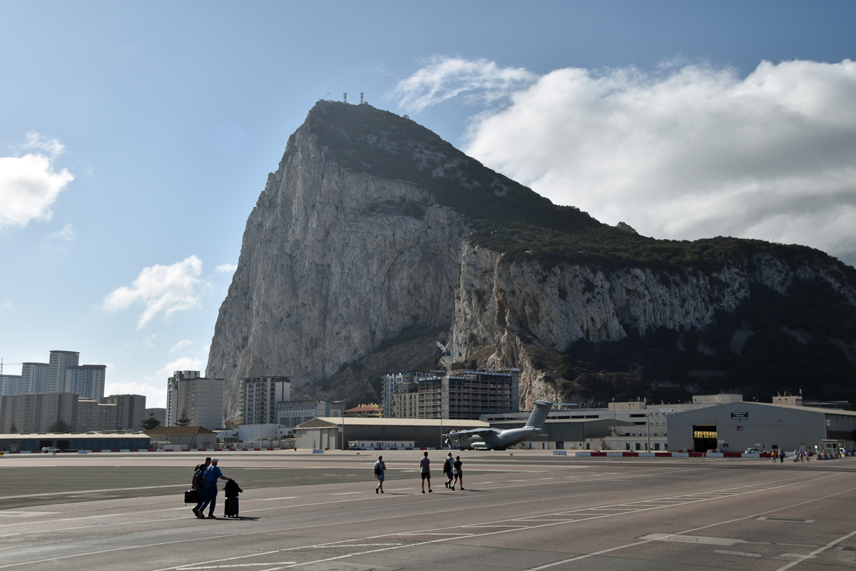 People walk across an airport runway with the Rock of Gibraltar towering in the background under a partly cloudy sky. An airplane is visible near the terminal and buildings line the base of the rock.