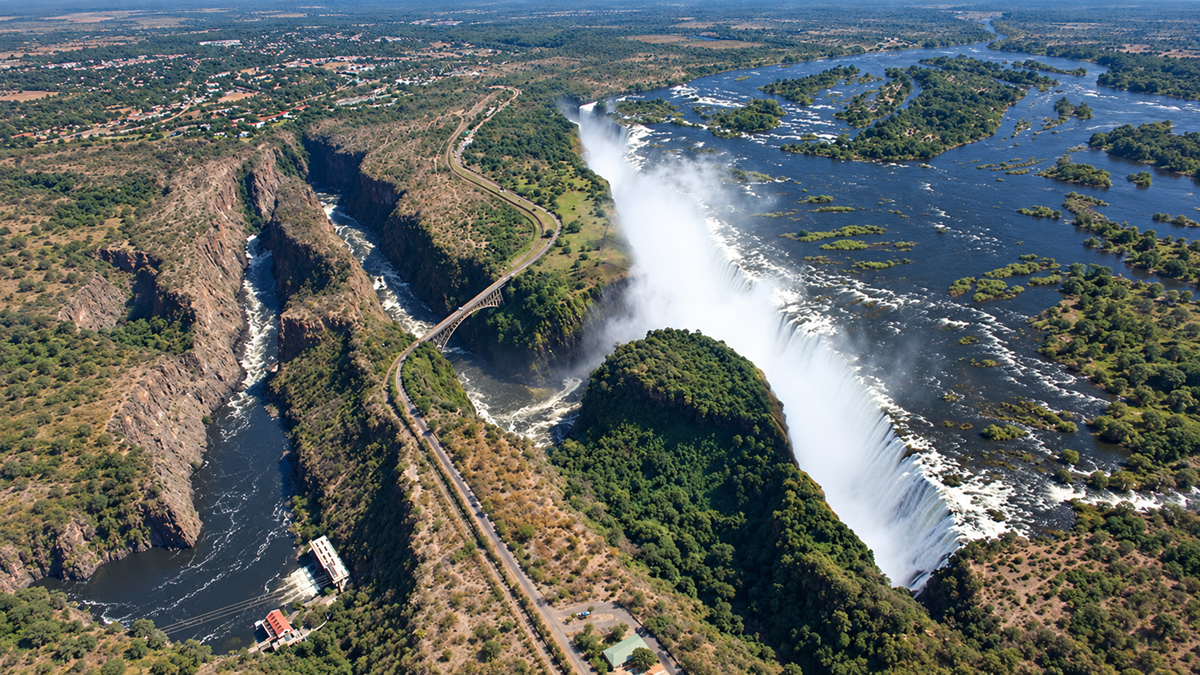 Aerial view of Victoria Falls with water cascading over cliffs, lush green islands, mist rising, and a river winding through a deep gorge. A bridge and roadway cross near the falls, with nearby buildings and distant landscape visible.