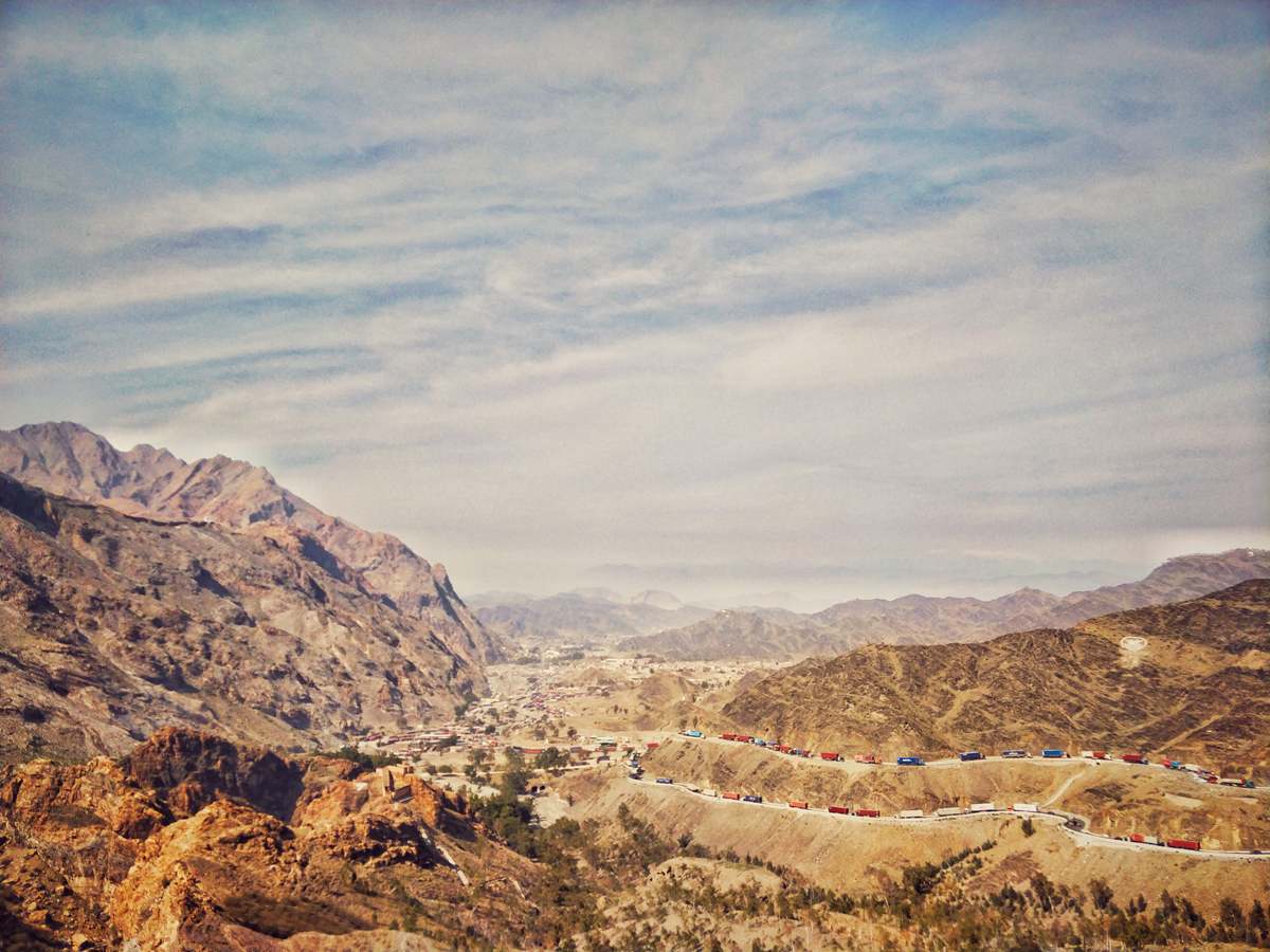 A winding mountain road with a long line of colorful trucks stretches through an arid, rugged landscape. Distant peaks and a hazy blue sky fill the background.