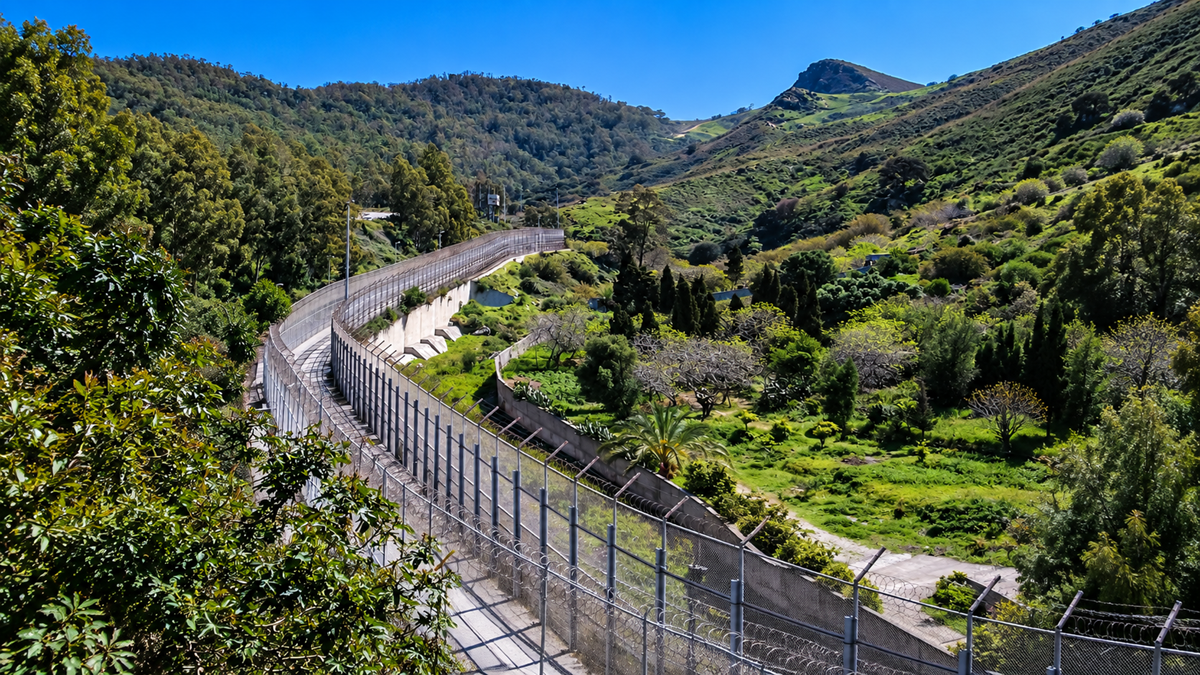 A tall metal fence winds through a lush, green hilly landscape, separating two areas. Trees and bushes line both sides under a clear, blue sky.
