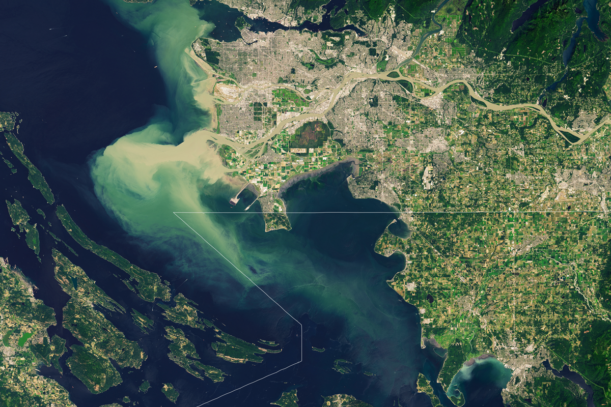 Satellite view of a coastal city with winding rivers, green land, and blue-green water mixing at the shoreline. Visible islands are below, and urban areas are concentrated near the top and left.