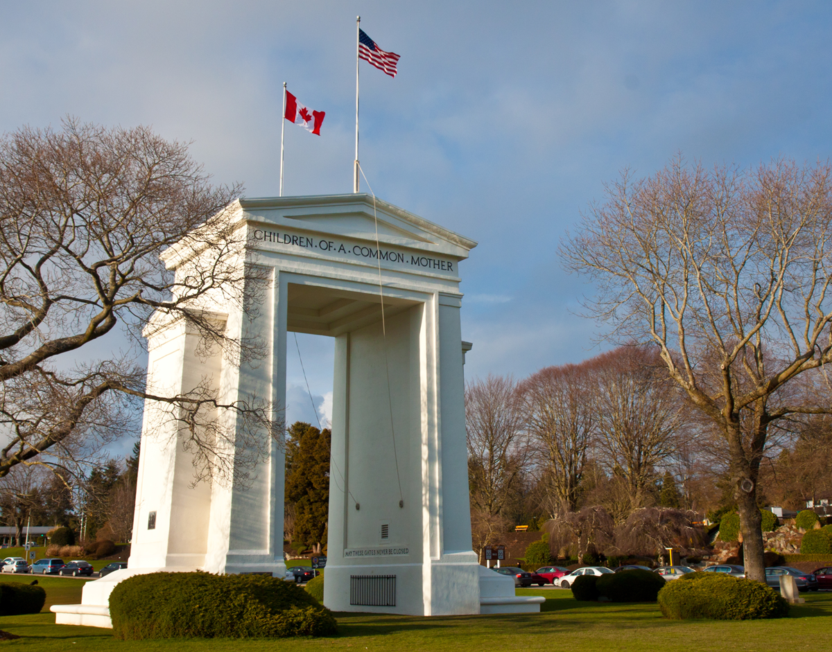 A white arch monument with U.S. and Canadian flags stands on a grassy area, surrounded by trees and parked cars. The arch reads, “CHILDREN OF A COMMON MOTHER.” The sky is partly cloudy.