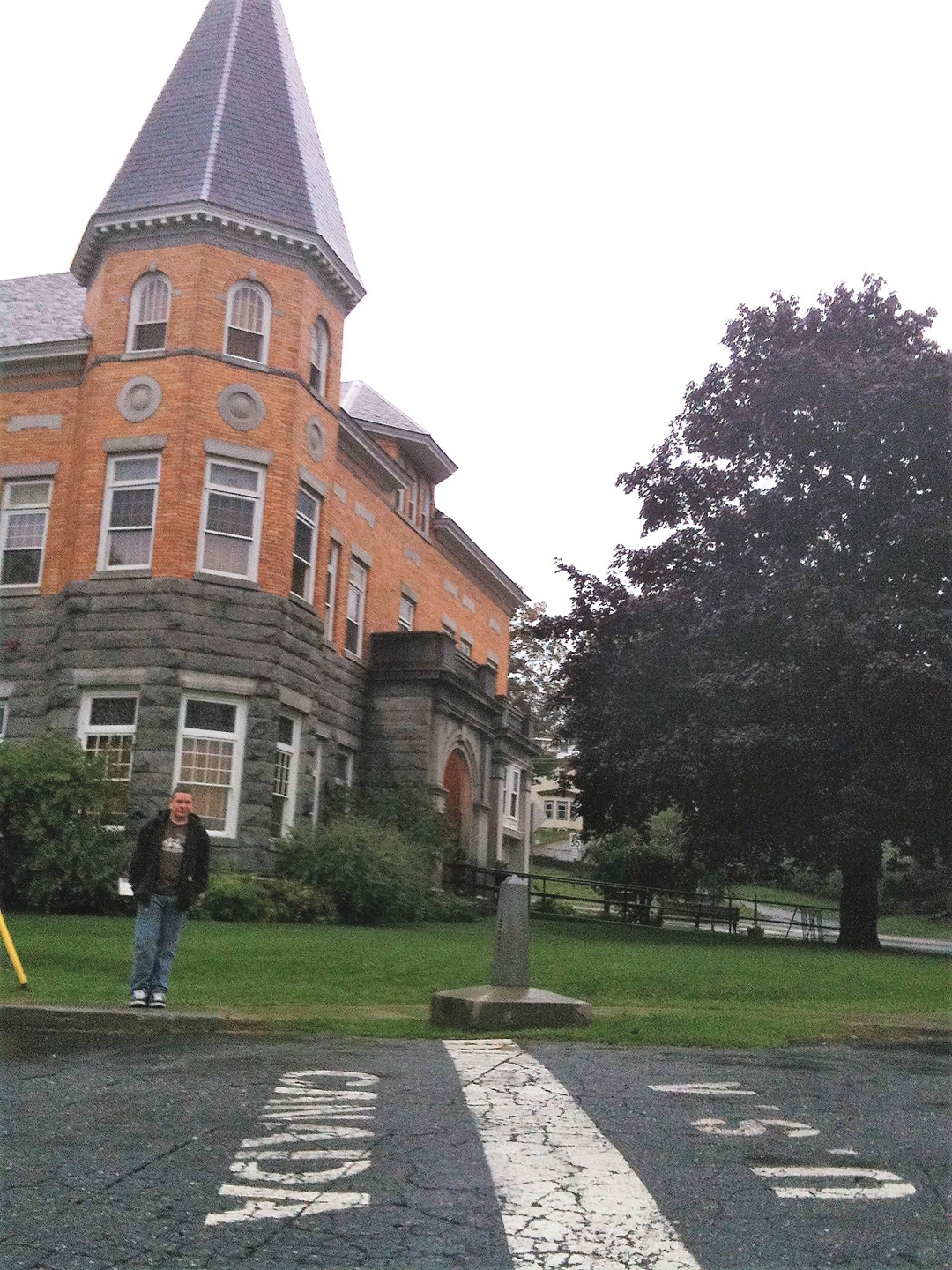 A person stands by a historic brick building near a line painted on the road marking the Canada-U.S.A. border, with a large tree and grass nearby.