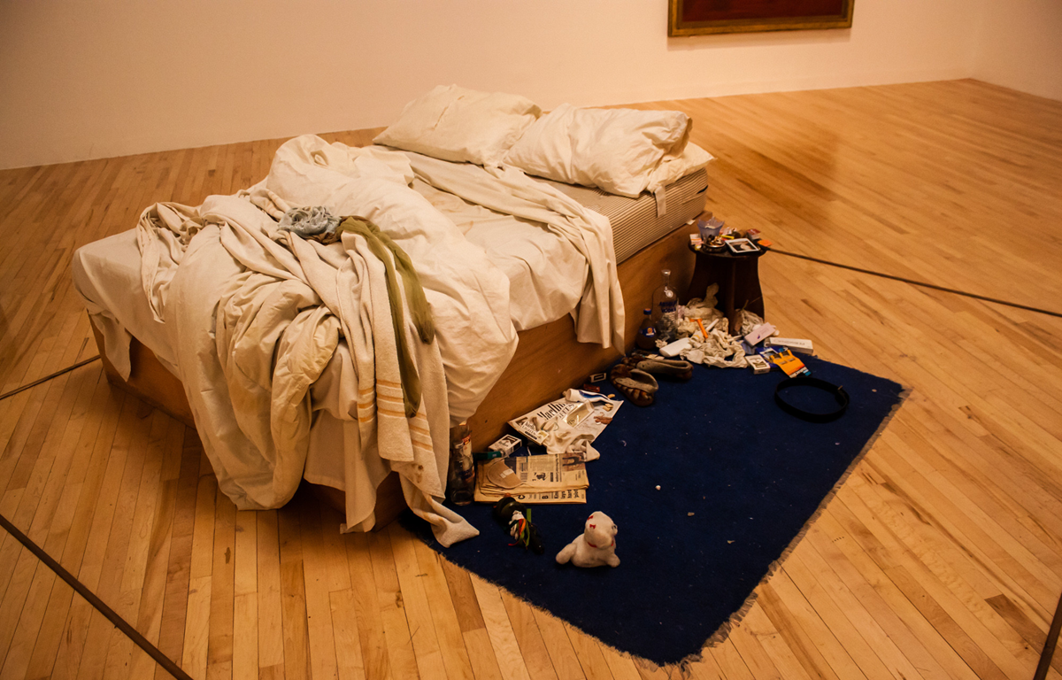 A messy unmade bed with white sheets sits on a blue rug, surrounded by scattered personal items, magazines, clothes, and a small stuffed toy, in a room with wooden floors and minimalist decor.
