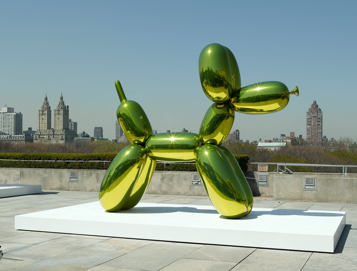 A large, shiny, green balloon dog sculpture sits on a white platform outdoors with a city skyline in the background under a clear sky.