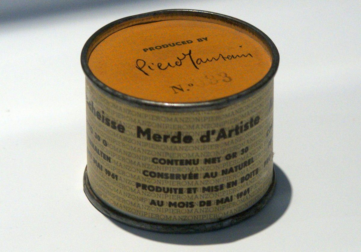 A small cylindrical can labeled “Merda d’Artista” with text in various languages and a handwritten signature on the gold lid, displayed on a white surface.