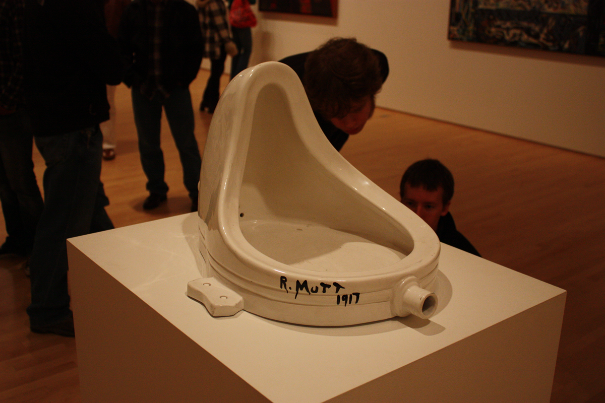 A white porcelain urinal titled “Fountain” signed “R. Mutt 1917” is displayed on a pedestal in an art gallery, with people observing in the background.