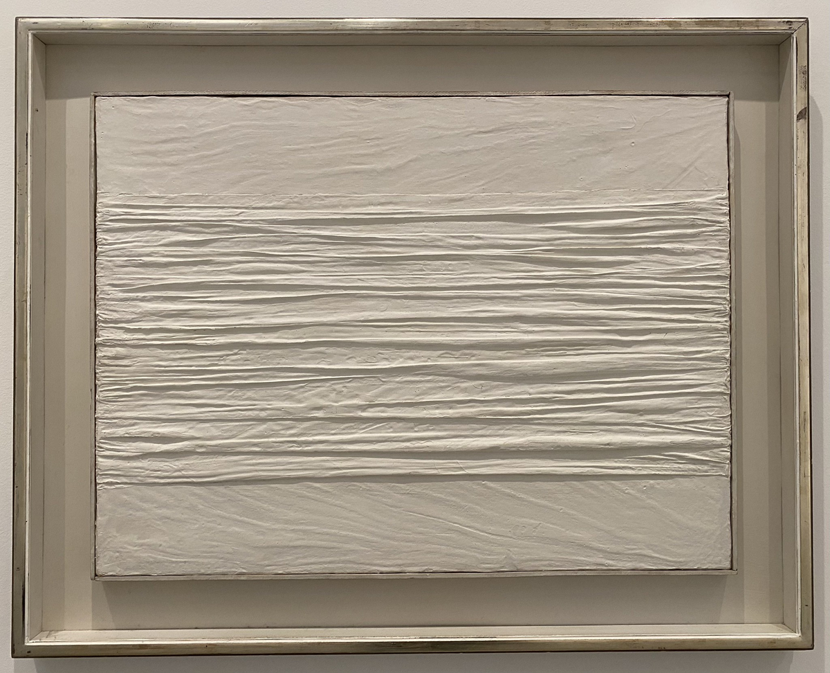 A framed artwork with a textured white surface, featuring horizontal ridges across the center, creating a wrinkled, fabric-like appearance. The piece has a minimalist and abstract style.