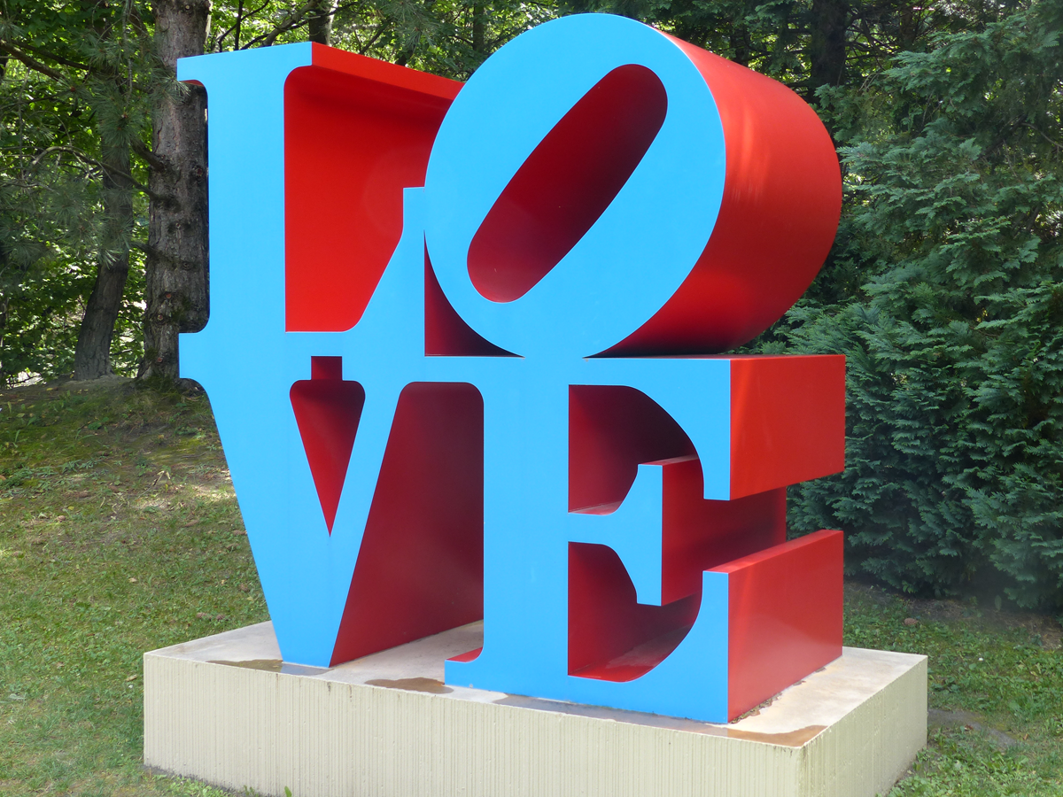 A large outdoor sculpture spells “LOVE” in blue letters with a red background, with the “O” tilted sideways, set on a concrete base surrounded by grass and trees.