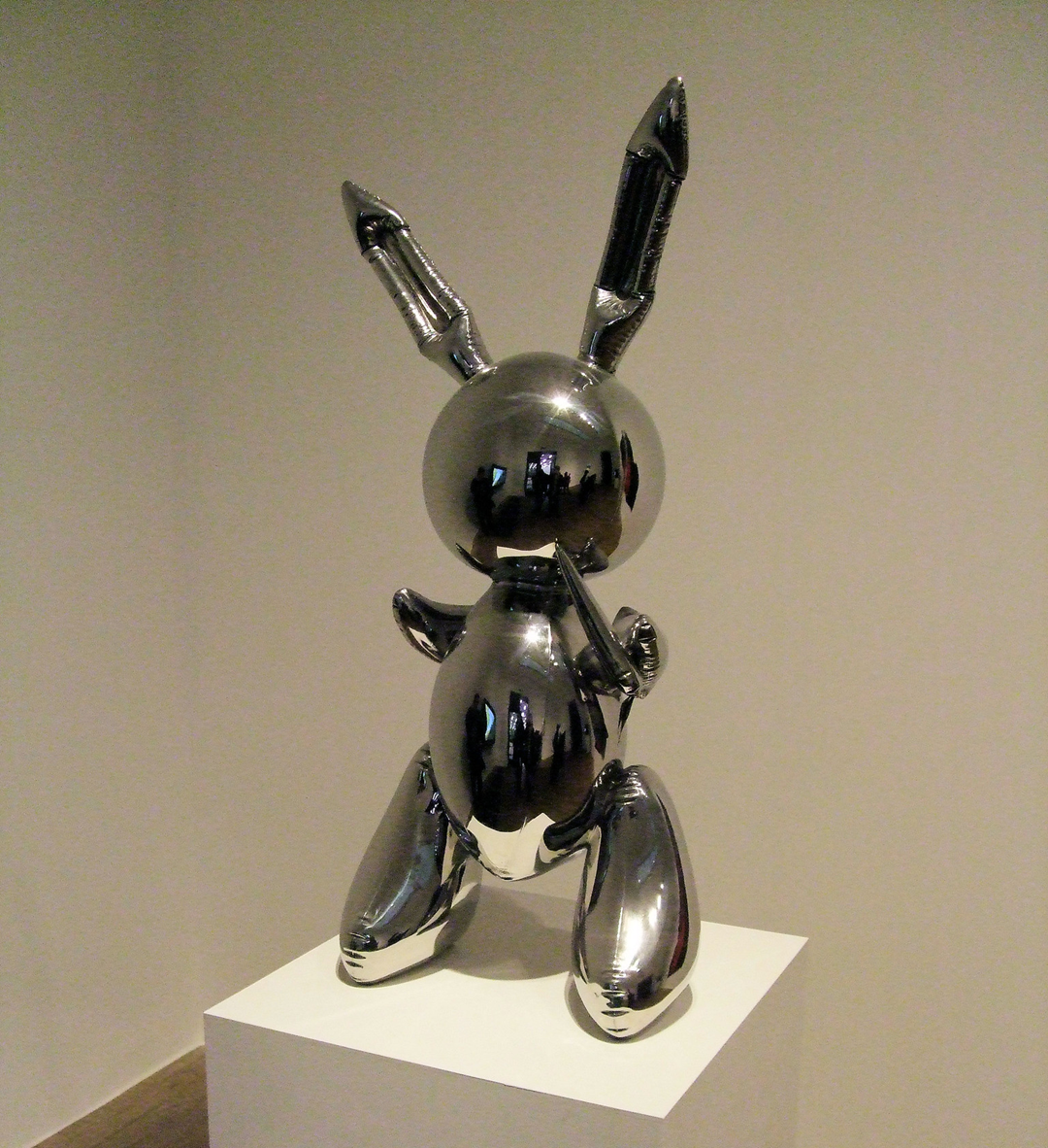 A shiny, silver balloon-like sculpture of a rabbit stands upright on a white pedestal against a plain background.