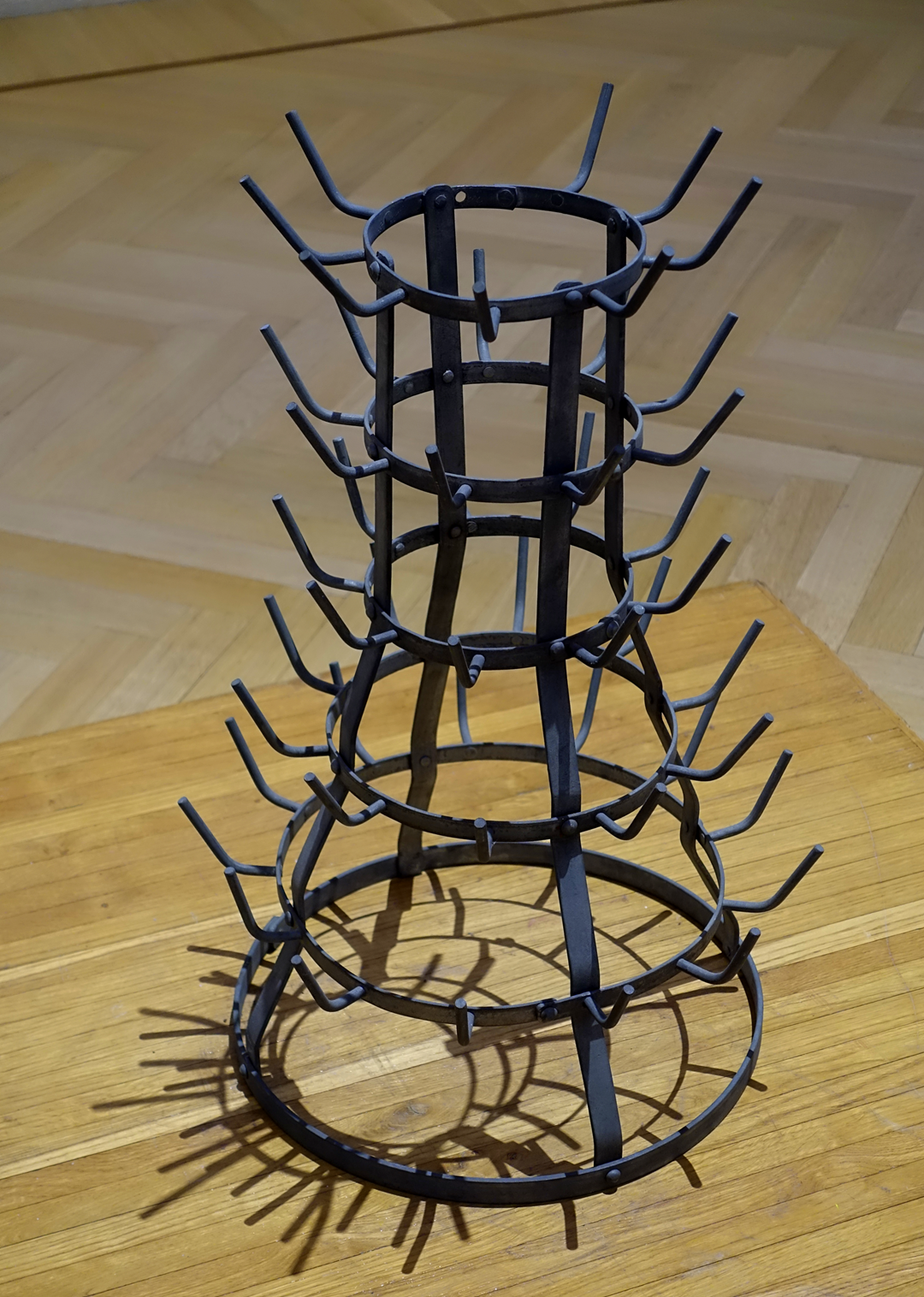 A black metal bottle drying rack with multiple rows of angled hooks, standing on a wooden floor with a parquet pattern.