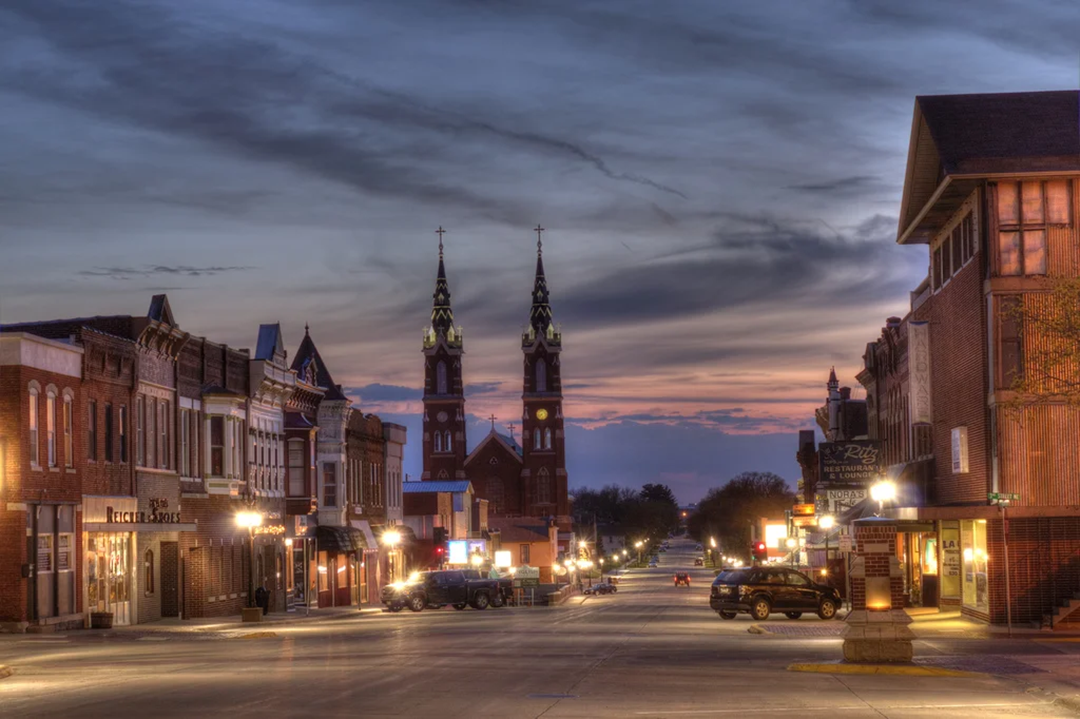 A quiet small-town street at dusk with historic brick buildings on both sides and a church with two tall spires in the distance, soft lights glowing from shops and street lamps.