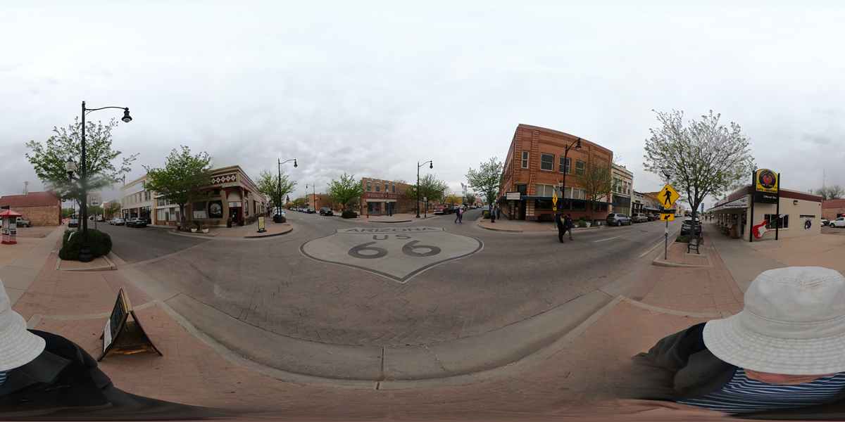 A 360-degree street view of downtown with the Route 66 shield painted on the road, brick buildings, trees, cloudy sky, and people walking. A person in a hat is partially visible at the bottom edge.