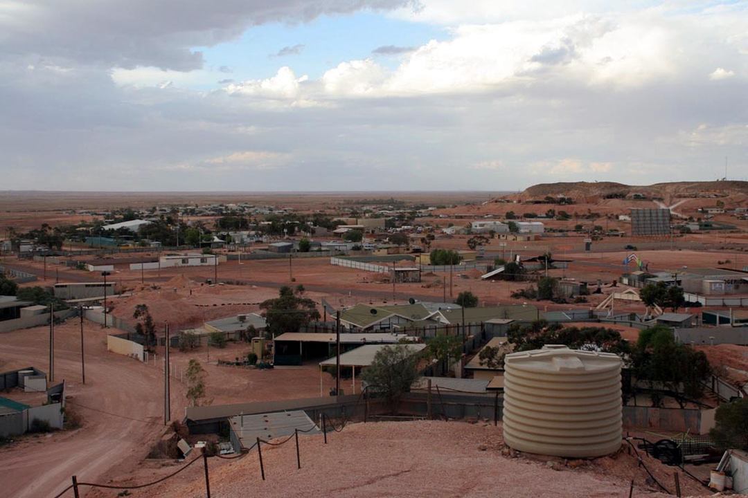 A view of a small desert town with reddish soil, scattered houses, a large water tank in the foreground, and a cloudy sky overhead.