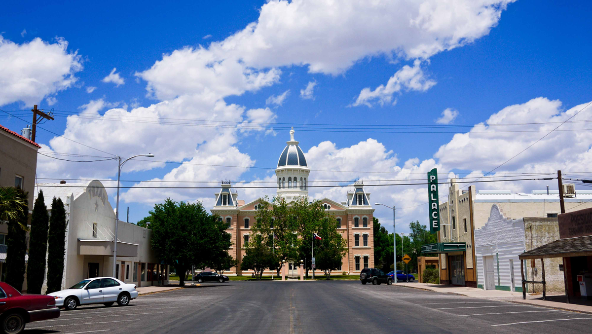A wide street leads to a historic courthouse with a central dome, flanked by trees and parked cars under a blue sky with scattered clouds. A green “Palace” sign is visible on the right.