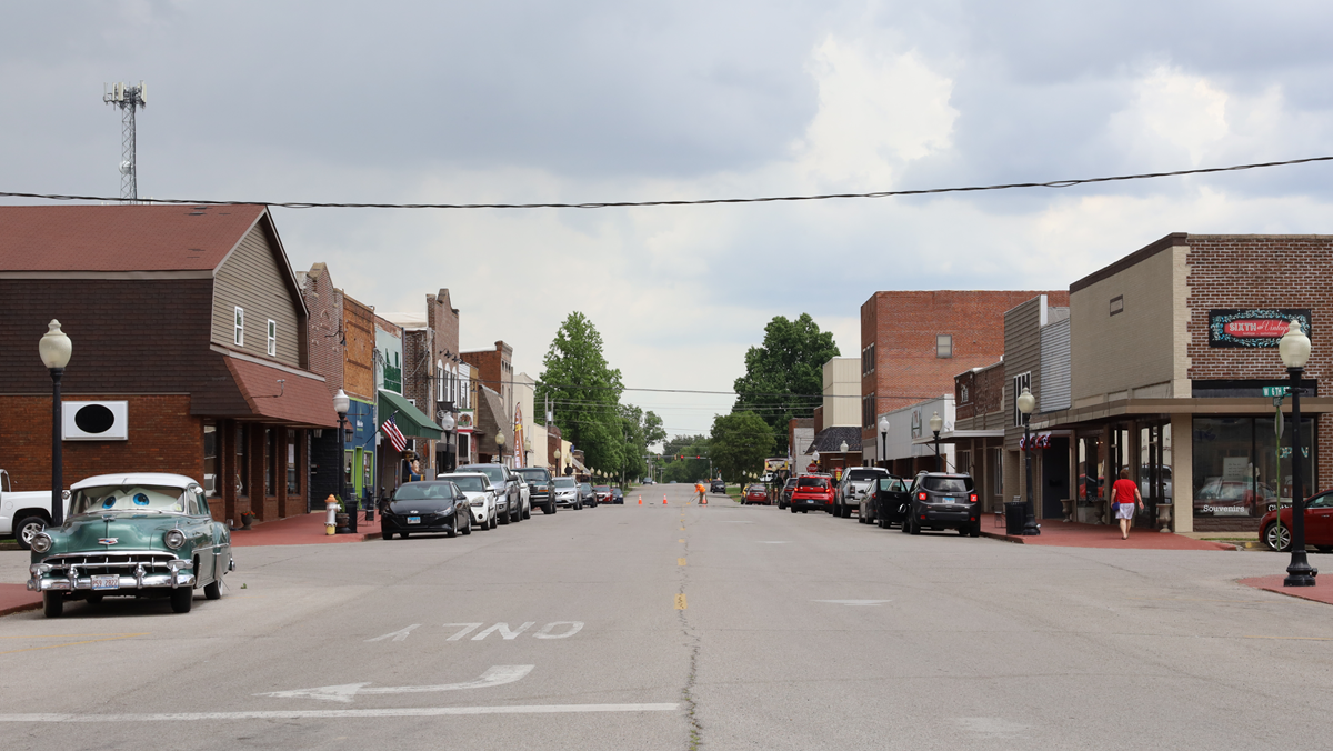 A small town main street with parked cars, brick buildings, and a vintage green car on the left. People walk on sidewalks under a cloudy sky.