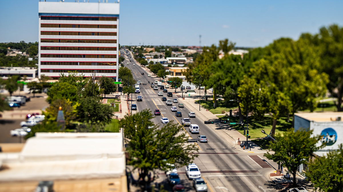 A tilt-shift, high-angle view of a multi-lane street in a town, featuring a prominent rectangular building with horizontal red and tan stripes on the left and a lush, blurry park with trees on the right, under a bright, clear blue sky.