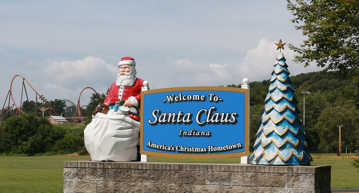 A “Welcome to Santa Claus, Indiana” sign is displayed with a Santa statue and a Christmas tree sculpture. A roller coaster and trees are visible in the background under a clear sky.