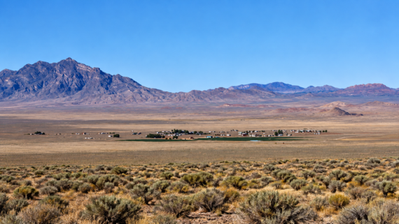 A small desert town sits in a vast, dry plain with sparse shrubs, backed by rugged, tall mountains under a clear blue sky.