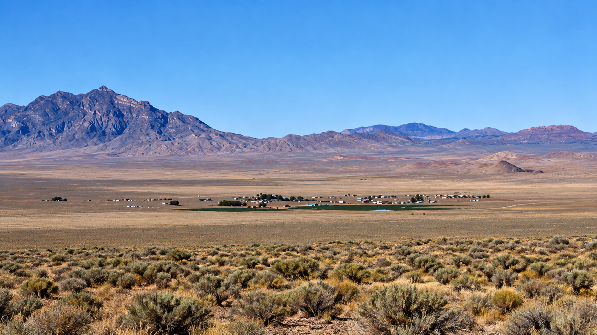 A small desert town sits in a vast, dry plain with sparse shrubs, backed by rugged, tall mountains under a clear blue sky.