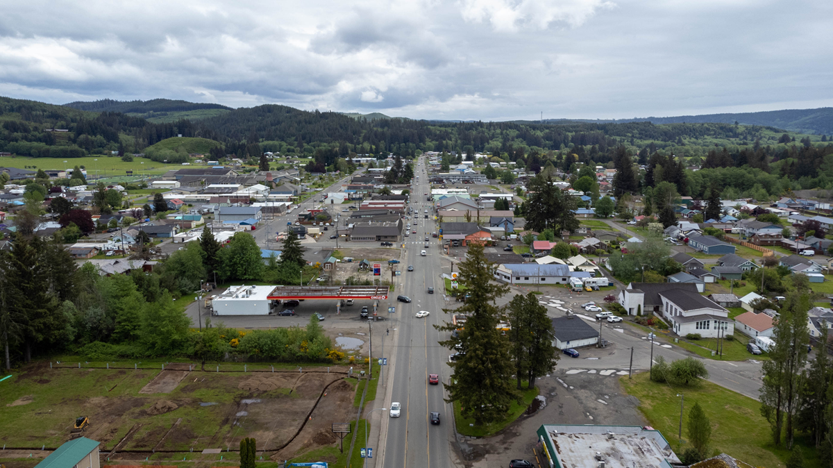 Aerial view of a small town with houses, trees, and hills in the distance under a cloudy sky. A main road runs through the center with cars and buildings along both sides.