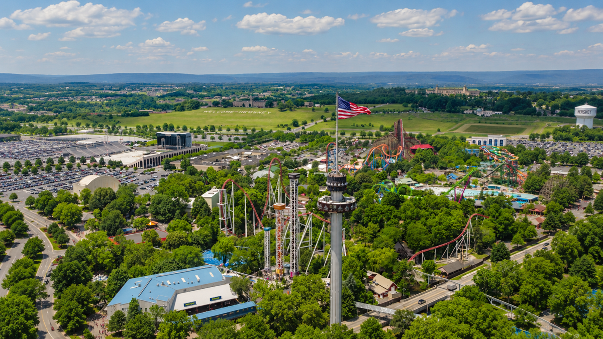 Aerial view of an amusement park with roller coasters, lush green trees, and a tall tower flying an American flag, surrounded by parking lots and open fields under a partly cloudy sky.