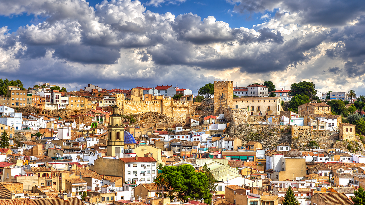 Colorful hillside town with tightly packed houses, historic stone castle walls, and towers under a dramatic cloudy sky. Lush trees dot the scene, adding greenery to the urban and ancient architecture mix.