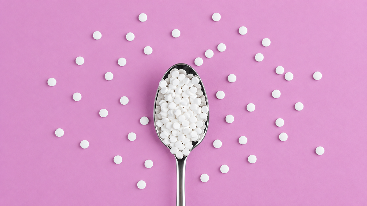 A metal spoon filled with small white round tablets, with more tablets scattered around it on a pink background.