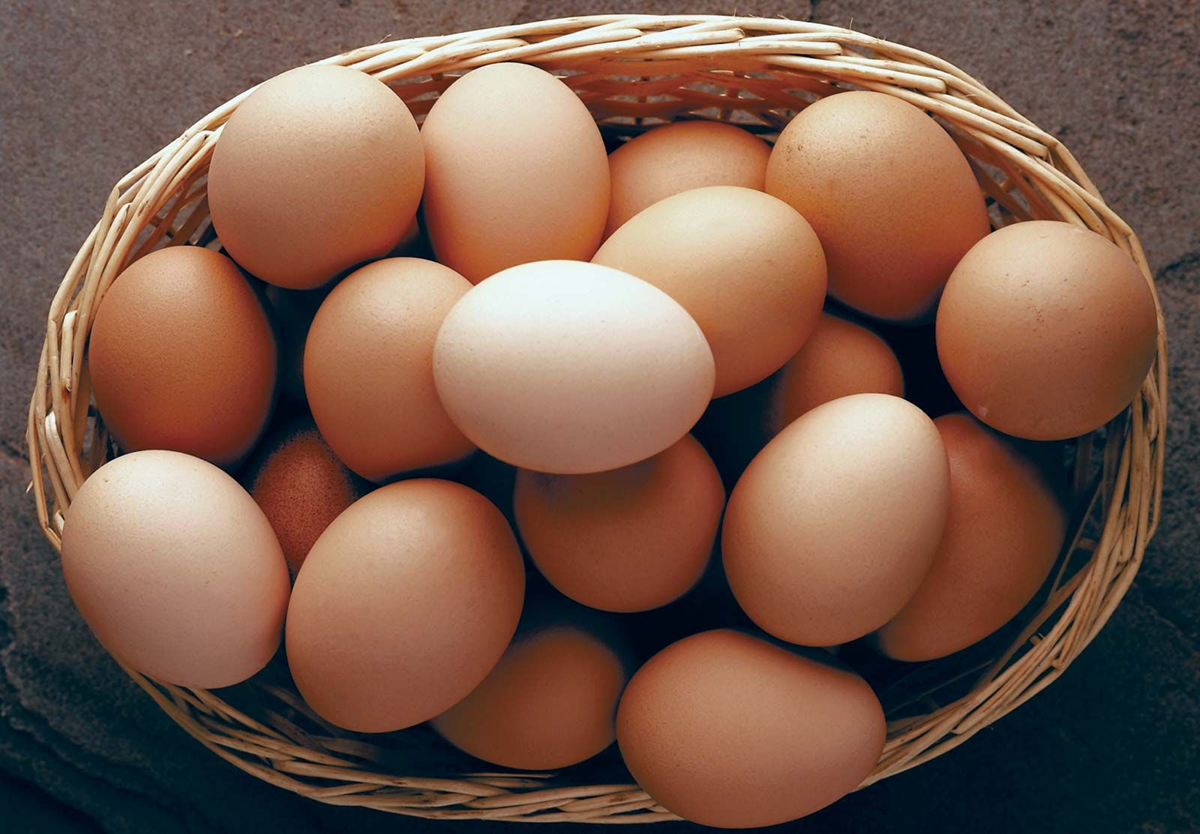 A wicker basket filled with several light brown eggs, placed on a dark, textured surface.