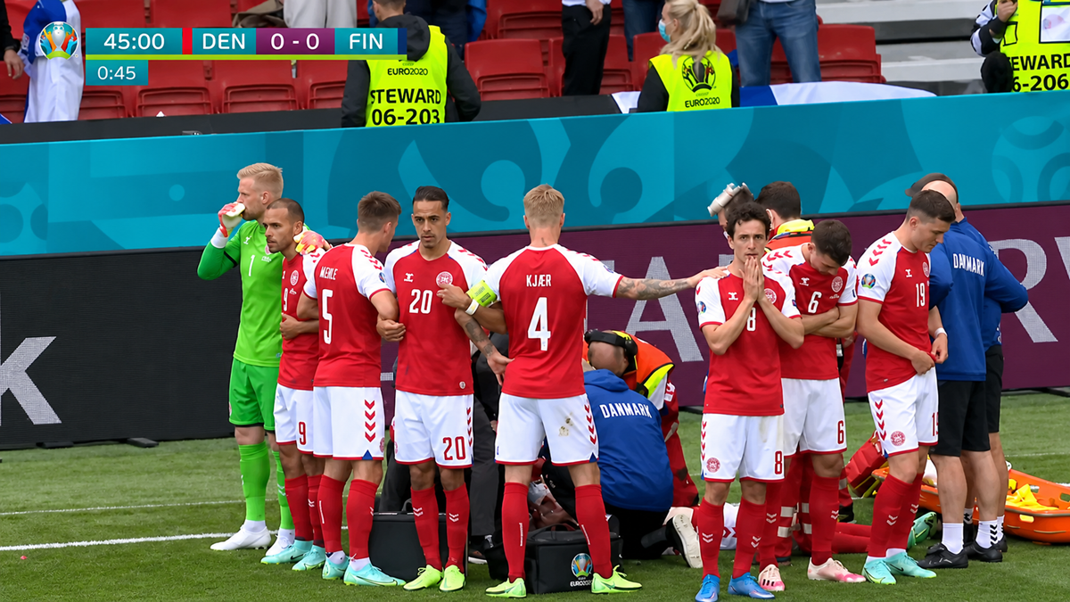 Danish soccer players huddle around a teammate receiving medical attention on the field during a match against Finland. Anxious expressions and concern are visible; staff and stewards are nearby. Scoreboard shows 0-0 at 45 minutes.