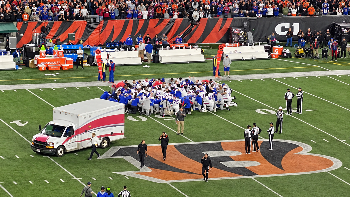 A football team kneels in a circle around a player on the field near an ambulance, with referees and staff nearby, as a crowd watches from the stands.