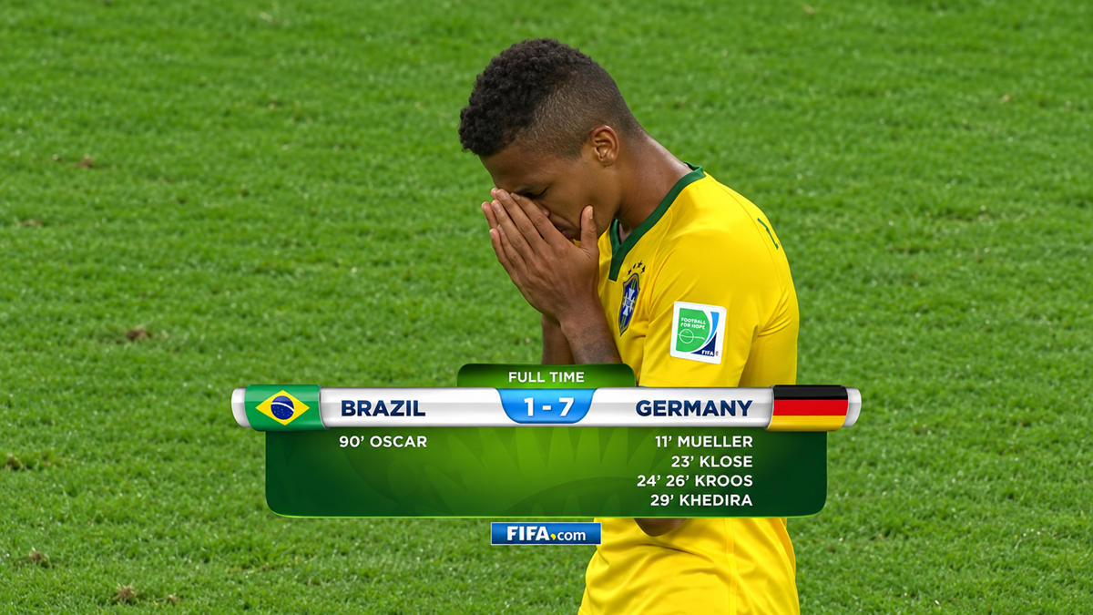 A Brazilian soccer player covers his face in distress after a 1-7 loss to Germany, as the scoreboard and goal scorers are displayed on screen over a grassy field.