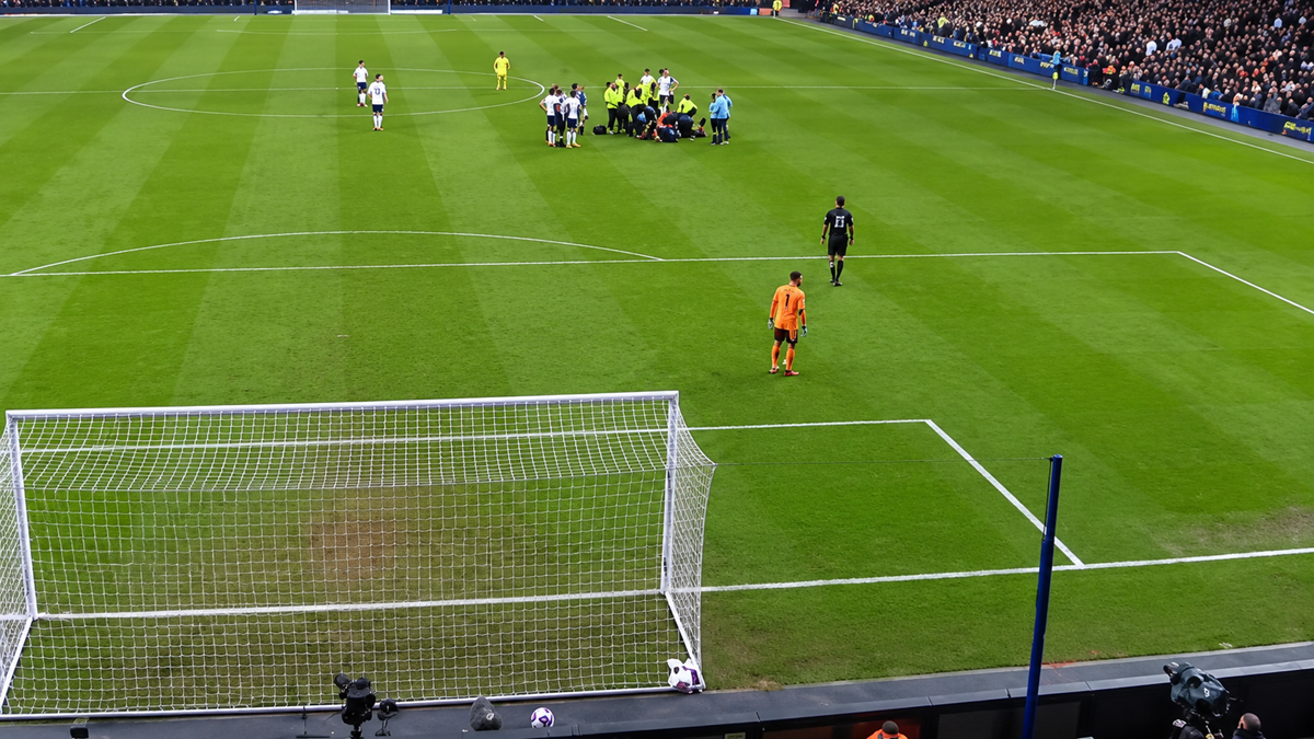 A soccer match in a stadium; players gather near the penalty area while a goalkeeper in orange stands by the goal. Fans fill the stands, watching the action unfold on the field.