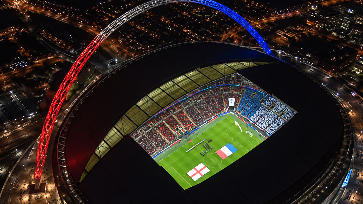 Aerial view of a lit stadium at night with England and France flags on the field. The stadium’s arch is illuminated red, white, and blue.