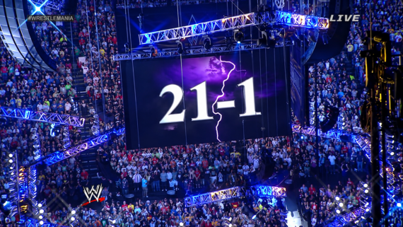 A large arena crowd watches as a screen displays “21-1” with a lightning bolt, under blue and purple lighting. The WWE logo and “WRESTLEMANIA” hashtag are visible.