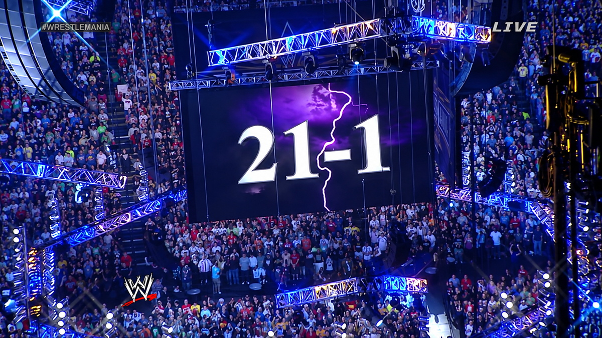 A large arena crowd watches as a screen displays “21-1” with a lightning bolt, under blue and purple lighting. The WWE logo and “WRESTLEMANIA” hashtag are visible.