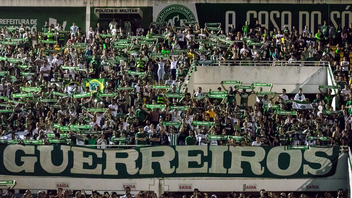 A large crowd of soccer fans holds green scarves and banners in a stadium. A large sign reading “GUERREIROS” is displayed at the front. Many people are wearing green and white.