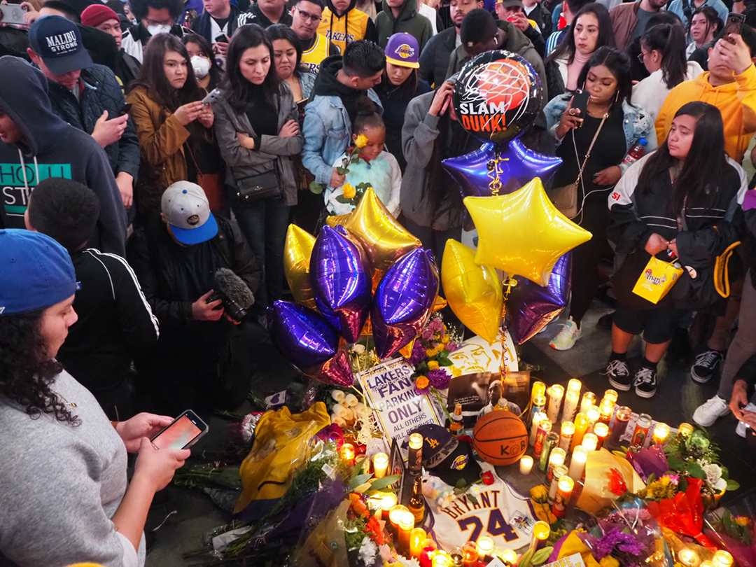 A crowd gathers around a memorial with candles, flowers, balloons, and basketball items, paying tribute to Kobe Bryant. Many people look emotional and somber, some taking photos or holding Lakers memorabilia.