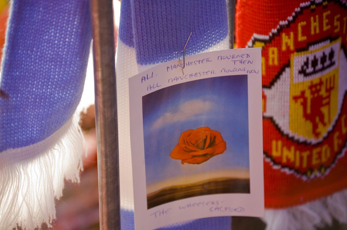 A photograph of a rose is attached to blue and red Manchester United scarves. A handwritten note reads, “All Manchester mourned then, all Manchester mourns now.” Sunlight illuminates the scarves and card.
