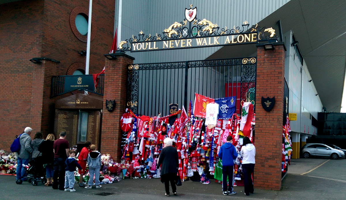 People stand by the Shankly Gates at Liverpool FC’s stadium, decorated with scarves, flowers, and tributes beneath the “You’ll Never Walk Alone” sign, honoring the Hillsborough disaster victims.