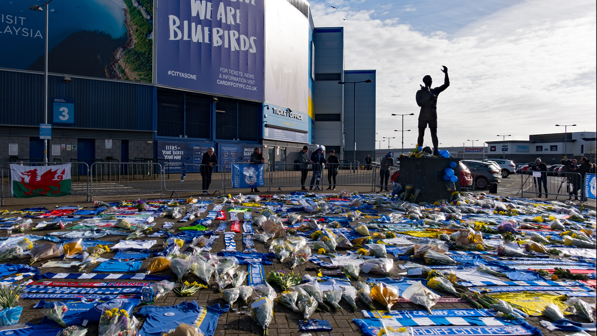 Flowers, scarves, and tributes cover the ground before a stadium statue, while people stand nearby. A large sign reads “We Are Bluebirds.” The scene honors someone significant to the community.